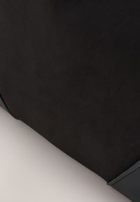 Close-up of a black fabric and leather item, showing texture and stitching details on a smooth light background.