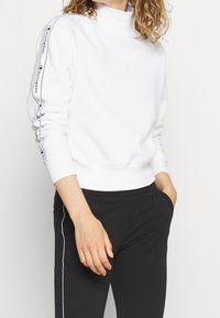 Felpa bianca con collo alto e logo nero sulle maniche, abbinata a pantaloni neri con una striscia bianca laterale. Materiale morbido e vestibilità comoda.
