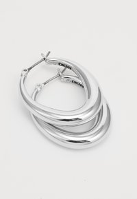 Une paire de boucles d'oreilles créoles en argent brillant, avec un design épais et arrondi et des fermoirs à crochet sécurisés, sur un fond blanc uni.