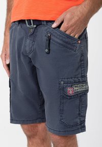Blue cotton cargo shorts with multi-pocket design, zippered and flap pockets, decorative stitching, and a logo patch on the side.