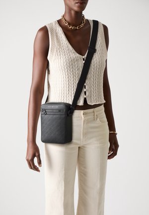 Frau trägt eine ärmellose cremefarbene Strickweste und cremefarbene Hose, sie trägt eine schwarze Umhängetasche mit strukturiertem Muster und Reißverschlusstasche.