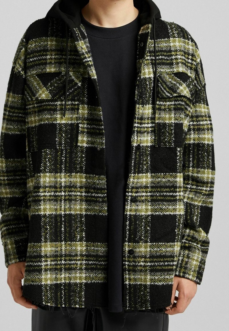 Černozelená kostkovaná bunda s texturovanou látkou, obsahující dvě náprsní kapsy, kapuci a mírně třepící se lem. Viditelná černá spodní košile.