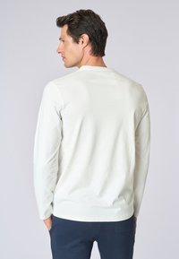 Camicia a maniche lunghe color beige chiaro, con collo rotondo e una texture morbida. Presenta un design semplice e un orlo rifinito. Indossata con pantaloni scuri.