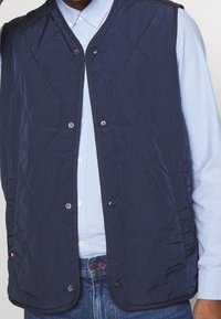 Marineblau gesteppte Weste mit Rautenmuster, ausgestattet mit Druckknopfverschlüssen und einer glatten Oberfläche. Getragen über einem hellblauen Hemd mit Kragen.