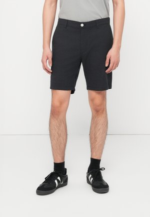 Mand iført sorte shorts med knapper, sorte sokker og sorte sneakers med hvide striber, stående mod en ensfarvet hvid baggrund.
