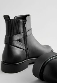 Bottines en cuir noir avec des détails en élastique côtelé, fermeture à glissière sur le côté et talon épais. Fini lisse avec des accents de coutures contrastées.