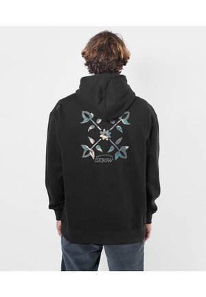 Personne aux cheveux courts et bruns portant un sweat à capuche noir avec un motif floral de tiges croisées et le mot OXBOW dans le dos.