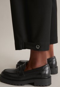 Schwarze, maßgeschneiderte Hose mit umgeschlagenem Saum und Knopfdetails, kombiniert mit schwarzen Mokassins mit einer klobigen Gummisohle und einer glatten Lederoberfläche.