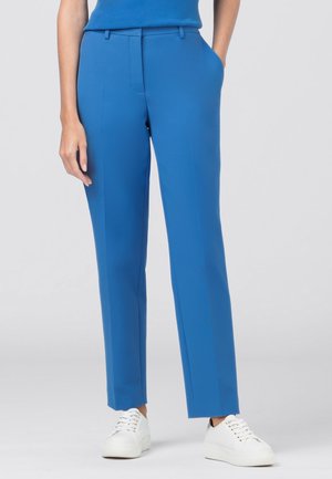 Vrouw die felblauwe rechte broek en witte sneakers draagt, staand met één hand in de zak tegen een effen achtergrond.