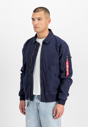 Man wearing navy blue bomber jacket with red tag on sleeve, white t-shirt, and light blue jeans, standing against a plain white background.