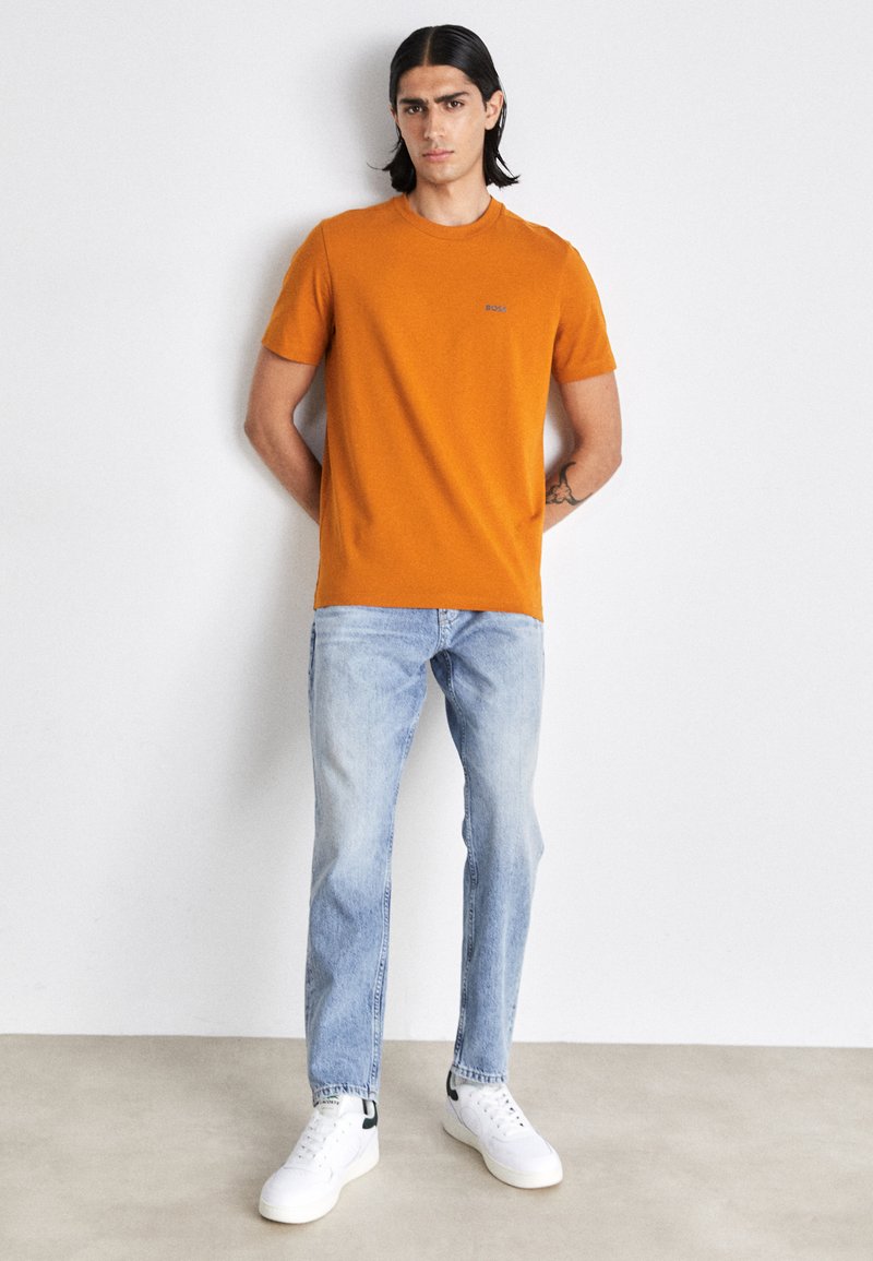 Maglietta arancione a maniche corte con un logo blu, abbinata a jeans azzurri e sneakers bianche, su uno sfondo neutro.