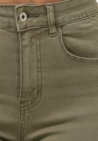 Los jeans de mezclilla verde oliva presentan un diseño clásico de cinco bolsillos, costuras visibles, un cierre de botón de metal y una textura suave.