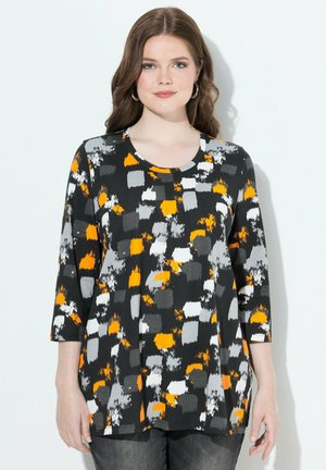 Vrouw in een zwarte top met grijze, witte en oranje abstracte vierkante patronen en driekwartmouwen, staande tegen een witte achtergrond.