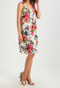Vestido sin mangas con estampado floral que presenta un fondo blanco, flores vibrantes en tonos rosa y verde, y un corte holgado. Sandalias de tacón alto con tiras en color plateado.