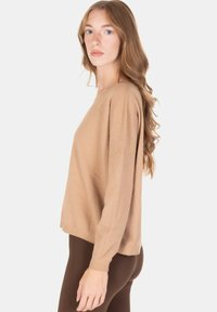 Donna con capelli lunghi e mossi, indossa un maglione beige e pantaloni marrone scuro, in posizione laterale su sfondo bianco.