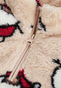 Chlpatá béžová fleece látka s červenými, čiernymi a bielymi vzormi; má krémový zips na prednej strane na zapínanie.
