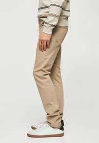 Persona vestida con pantalones slim-fit beige, zapatillas blancas con suelas marrones y un suéter beige con rayas negras y marrones, de pie de lado.