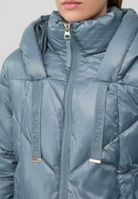 Veste matelassée bleue avec col haut, dotée d'une fermeture éclair intégrale et de sangles avec logo. Le tissu a une texture lisse et brillante avec un matelassage en forme de diamant.