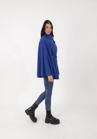Capa azul a la altura del torso con cuello alto, que presenta un cinturón negro. Combinada con jeans ajustados y botines negros gruesos. Textura suave.