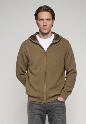 Homme aux cheveux châtain clair portant un sweat à capuche marron zippé sur un t-shirt blanc et un jean gris, debout avec une main dans la poche.