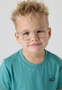 Vaikas dėvi skaidrius stačiakampius akinius ir melsvos spalvos trumpą rankovių marškinėlį su tamsiai mėlynu Levi's logotipu, turi trumpus, smailius plaukus.