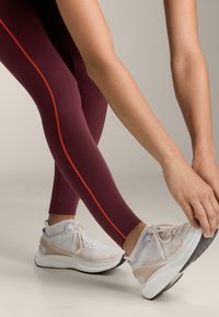 Bordové legíny s tenkým oranžovým pruhom na boku, kombinované s svetlošedými a bielymi bežeckými topánkami s béžovými akcentmi a štruktúrovanými podrážkami.