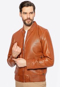 Giacca bomber in pelle marrone con polsini e colletto a coste, dotata di una zip frontale e una leggera lucentezza. Modello che fa gesti con le mani.