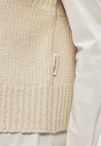 Suéter de malha em tom de creme, com tecido texturizado, barra canelada e etiqueta roxa da Calvin Klein na lateral. Posicionado ao lado de uma peça branca.