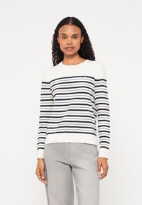 Stripet genser i krem og marineblå, med lange ermer, ribbestrikkede mansjetter, og en liten logo detalj på brystet. Myk strikketekstur.