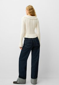 Pull en tricot blanc avec un col large, texture côtelée, associé à un jean large bleu marine au design classique à cinq poches.