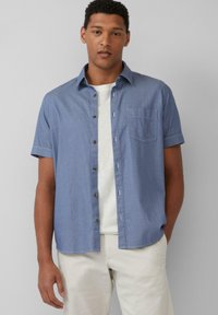 Koszula z krótkim rękawem w jasnoniebieskim kolorze, z tkaniny chambray, z kieszonką na piersi i brązowymi guzikami, noszona na białej koszulce.
