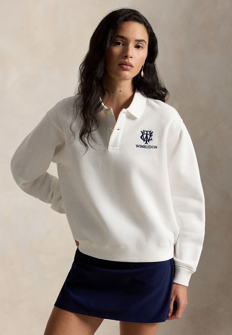 Femme aux longs cheveux foncés portant un sweat-shirt blanc avec le logo de Wimbledon et une jupe bleu foncé, debout devant un fond beige uni.