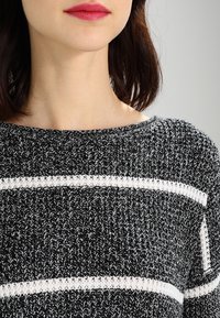 Mustapohjainen ja valkoinen neulepaita, jossa on leveä kaula-aukko ja teksturoitu kangas. Ominaisuuksina vuorottelevat kiinteät ja avoimet neuleraidat.