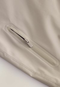 Țesătură impermeabilă bej, cu o textură netedă; dispune de un buzunar cu fermoar ascuns, cu un alunecător și o bandă asortate, amplasat pe lateral.