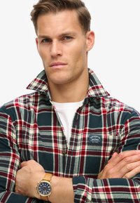 Rood en groen geruite flanellen shirt met een hoge kraag en een klein logo-patch, gedragen over een wit T-shirt; tan polshorloge zichtbaar.