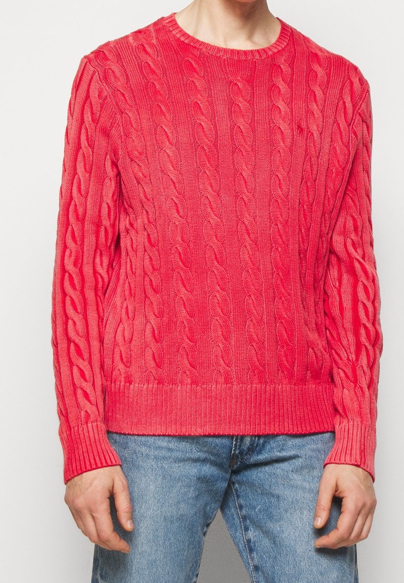 Maglione rosa a maglia cable con polsini e orlo a coste, caratterizzato da un motivo testurizzato. Indossato con jeans blu, che mettono in risalto una silhouette aderente.