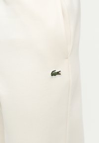 Κοντινή λήψη μπεζ παντελονιού με ένα μικρό πράσινο λογότυπο κροκοδείλου κεντημένο κοντά στην τσέπη.