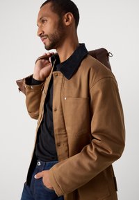 Hombre vestido con chaqueta marrón y cuello negro, pantalones vaqueros oscuros, llevando una bolsa de cuero marrón enrollada al hombro, posando frente a un fondo blanco.