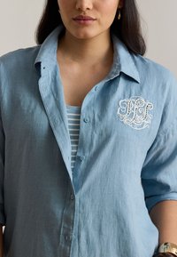 Vrouw draagt een lichtblauw linnen overhemd met opgerolde mouwen, een geborduurd monogram op de borst, een gestreept hemd eronder en gouden druppeloorbellen.