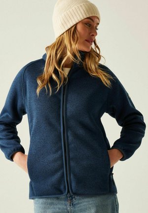 Vrouw met een crèmekleurige beanie en een navy fleecejas met rits en handen in de zakken, kijkt naar de zijkant tegen een effen achtergrond.