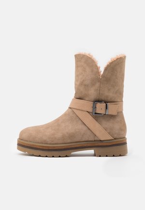 Bottine en suede beige avec une large ouverture, une doublure en peluche et une sangle décorative avec une boucle en argent. Semelle plate en caoutchouc texturé.