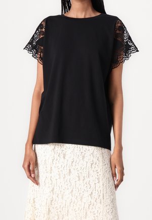 Sort kortærmet top med blonder på ærmerne, der har en afslappet pasform og glat tekstur. Båret over en cremefarvet blondekjole.