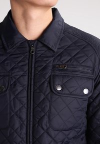 Marineblauwe gewatteerde jas met een rits aan de voorkant, drukknoopsluitingen, twee klepzakken en tekstuurelementen op de schouders. Bevat een kraag en een gladde stof.