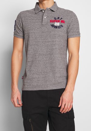 Homme portant un polo gris à manches courtes avec le texte « D.R.Y SUPER NO. 28 JAPAN ATH » et un pantalon cargo noir sur fond blanc.