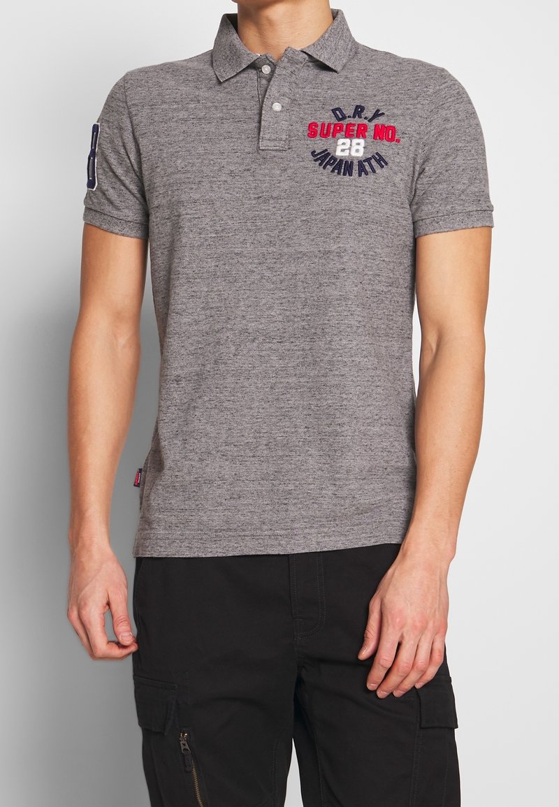 Homme portant un polo gris à manches courtes avec le texte « D.R.Y SUPER NO. 28 JAPAN ATH » et un pantalon cargo noir sur fond blanc.