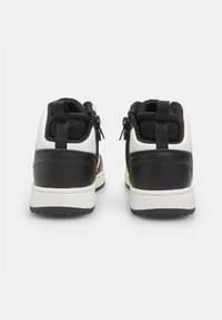 Zwart-witte sneakers met een bovenwerk van mesh, voorzien van een trekband aan de hiel en decoratieve accenten, geplaatst op een gestructureerde zwarte rubberen zool.