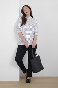 Chemise blanche à pois, manches longues, associée à un jean noir slim et des mocassins noirs brillants. Tient un sac fourre-tout noir, debout contre un mur.