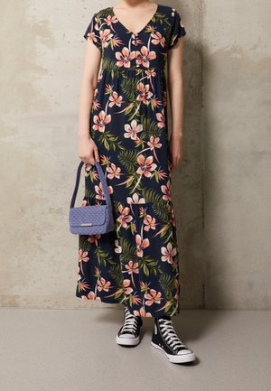 Kobieta ubrana w granatową, kwiatową sukienkę maxi, czarne wysokie trampki, trzymająca małą, fioletową torebkę o fakturze, stojąca przy beżowej ścianie.