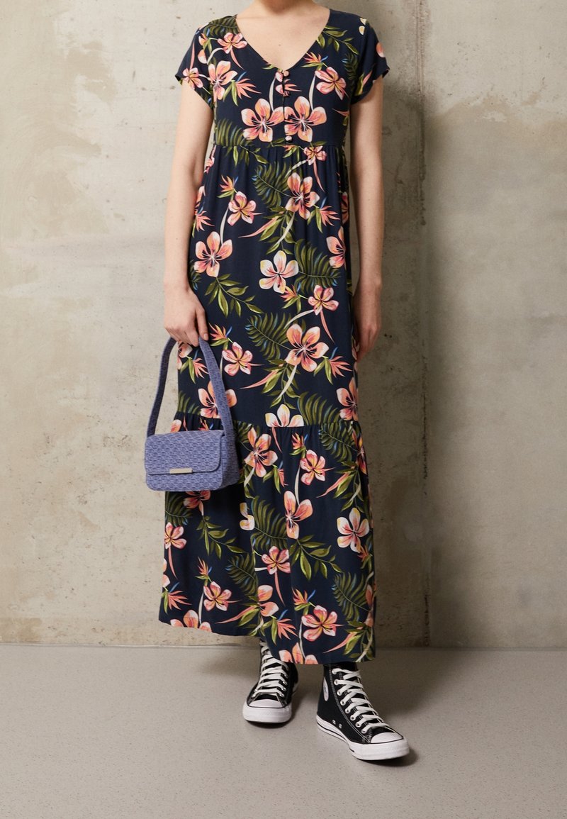 Femme portant une robe maxi à fleurs bleu marine, des baskets montantes noires, tenant un petit sac à main texturé violet près d'un mur beige.