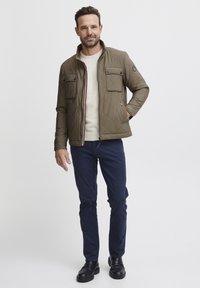 Quilted olive green jacket with two chest pockets and a striped zipper. Worn over a cream sweater and navy pants, paired with black shoes.